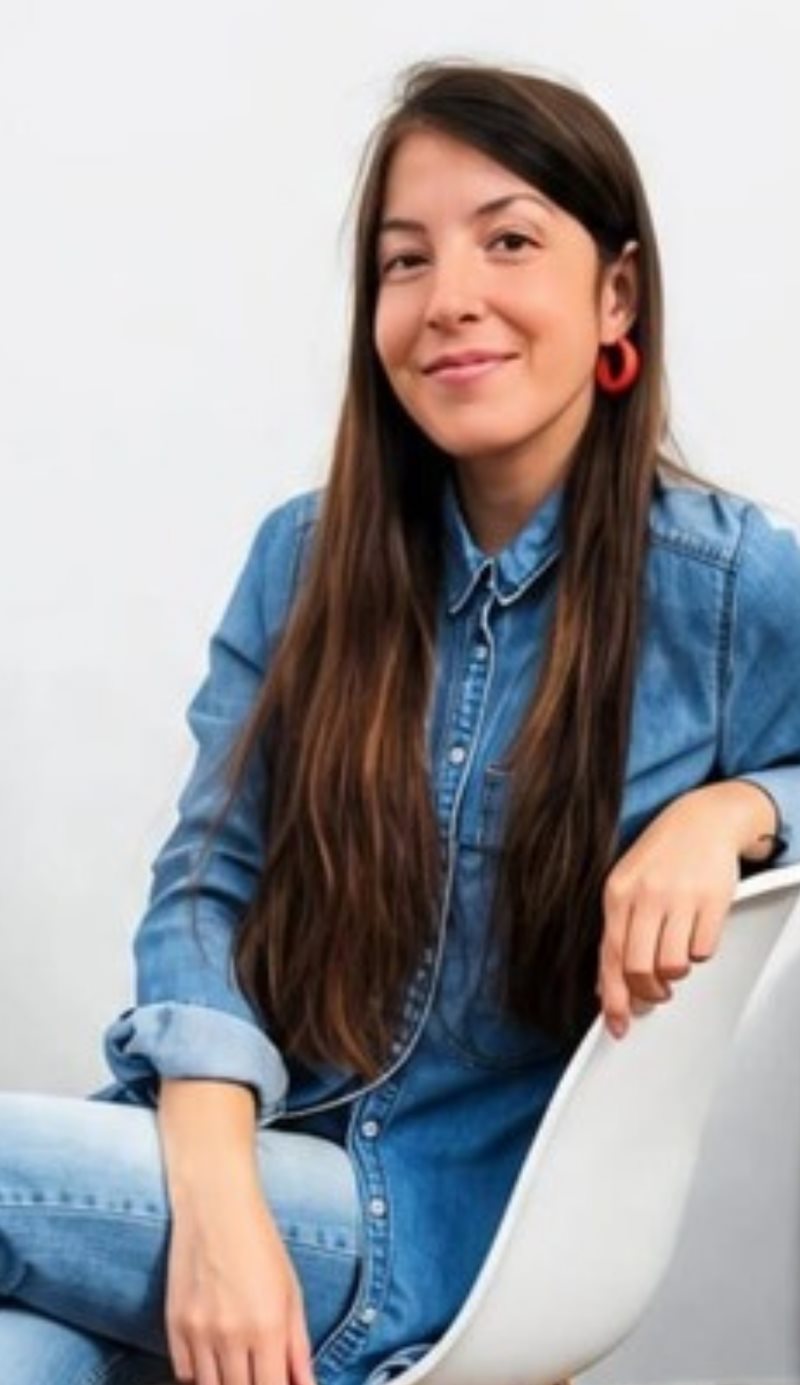 Dama sonriendo, sentada en una silla blanca, con camisa de jean